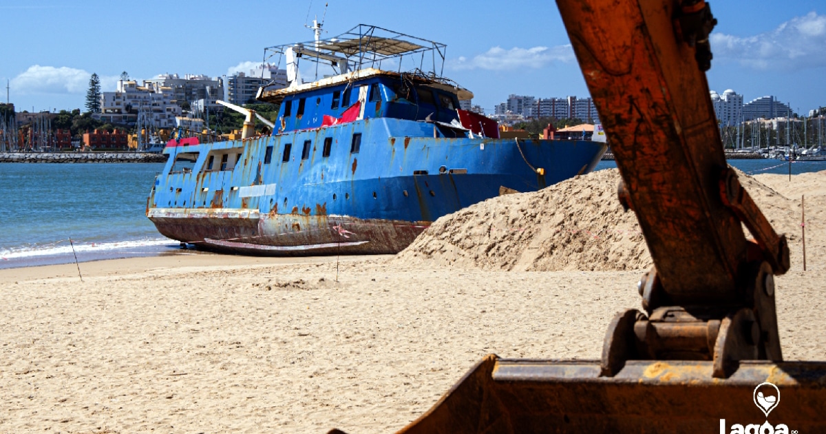 Barco encalhado na praia com escavadora em primeiro plano.